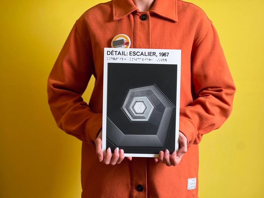 A person in an orange coat holds a tactile print titled “Détail: Escalier, 1967,” paired with a braille caption, showing a spiralling hexagonal staircase reduced to raised geometric layers.