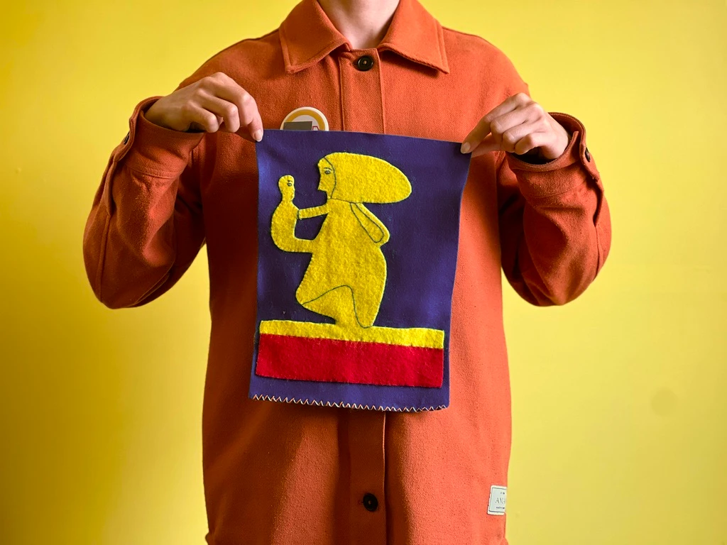 A person holds a felt textile piece appliquéd with a kneeling yellow figure on a purple ground above a red band — a soft, touchable rendering of a figure in motion.