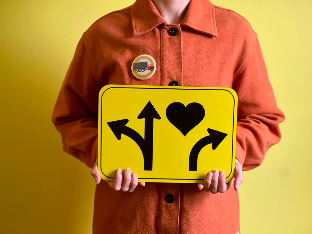 A person holds a bright yellow tactile wayfinding sign with raised black shapes: a forked arrow pointing left and up, a heart, and a curved arrow pointing right.