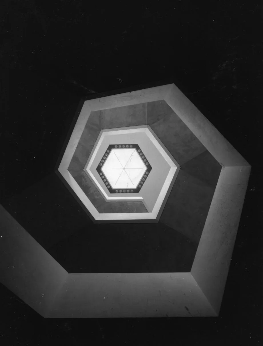 A colour photograph looking straight down through a spiralling hexagonal stairwell at the National Arts Centre, its nested hexagonal landings narrowing toward a bright skylight at the centre.