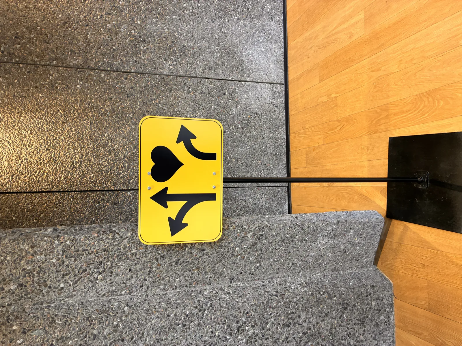 A yellow directional wayfinding sign mounted on a floor stand, showing a forked arrow pointing left and up, a heart, and a curved arrow pointing right, against a textured concrete wall.