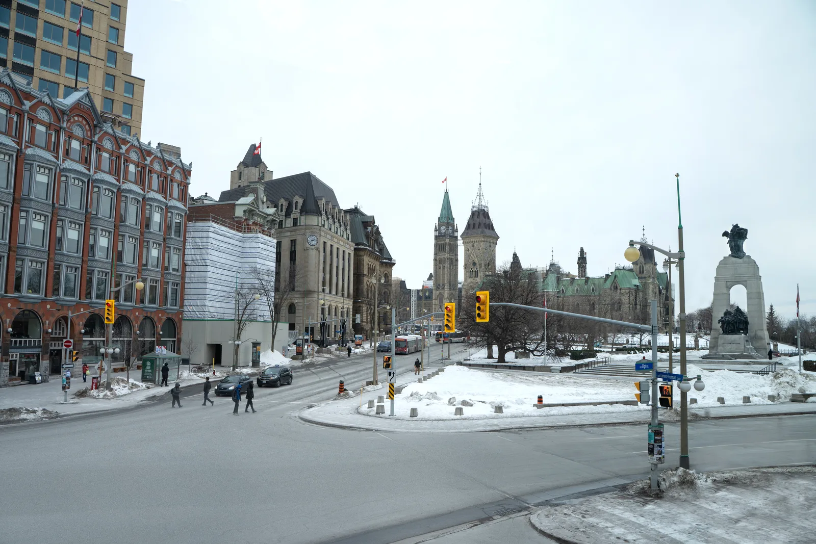 A wide street-level view of downtown Ottawa in winter, looking toward the Parliament Buildings’ Peace Tower and the National War Memorial, with pedestrians crossing a snow-lined intersection.