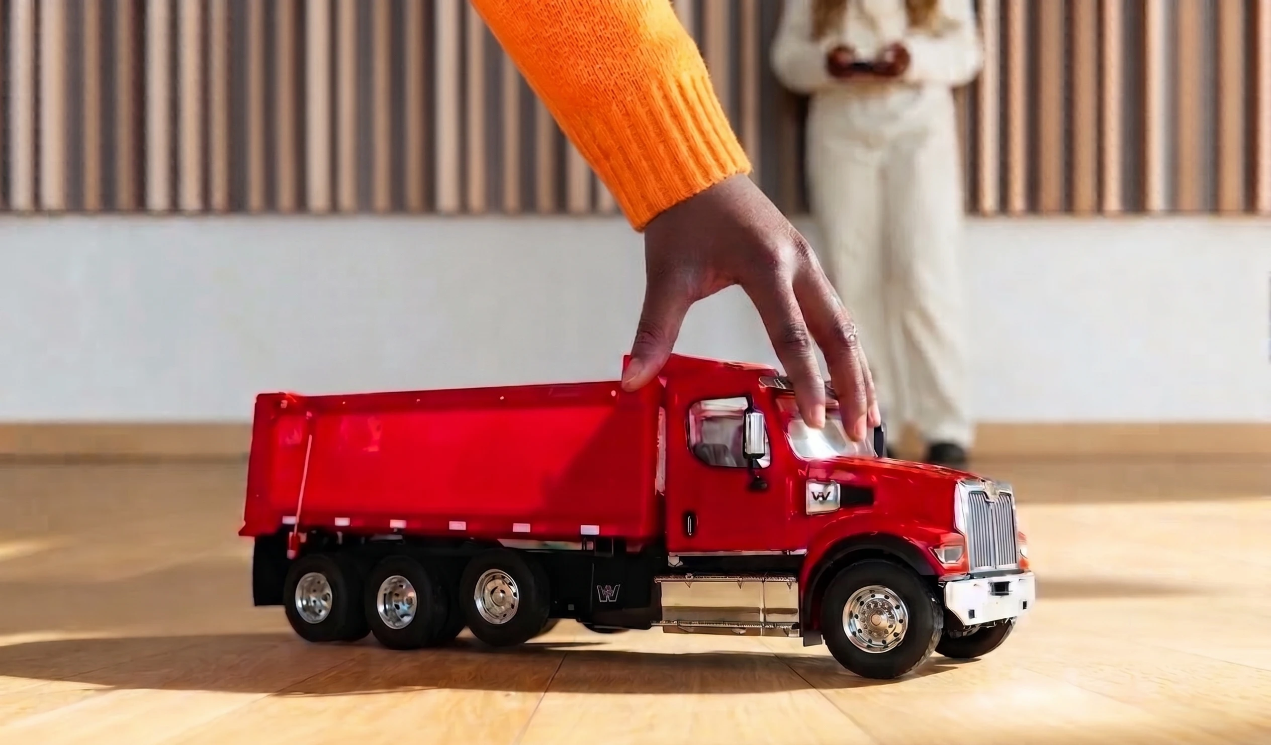 A hand reaches down and grips the roof of a bright red toy dump truck's cab as it sits on a hardwood floor, showing touch as a way of engaging with an object.